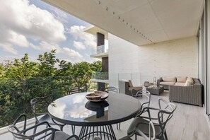 A modern outdoor dining area with a round table and chairs, a sofa, and a balcony with a view of trees and a building. - 3BR Playacar 3rd Floor - Rooftop Pool, Gym, Gated 24/7 (Playa del Carmen)