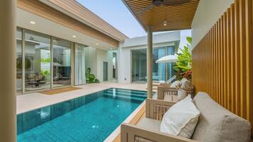 Un área de piscina moderna con una sala acristalada, una terraza de madera y un rincón de estar con cojines.