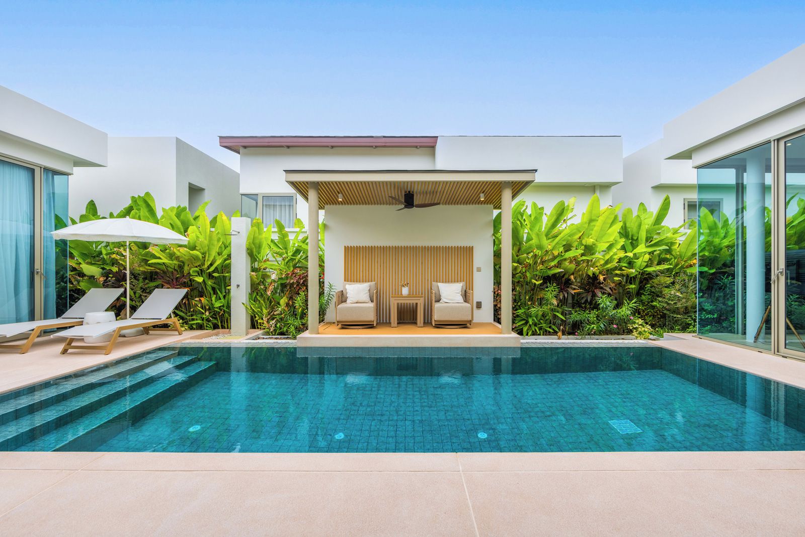 A modern pool area with lounge chairs, a seating area with cushions, and lush greenery.
