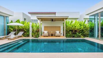 A modern pool area with lounge chairs, a seating area with cushions, and lush greenery.