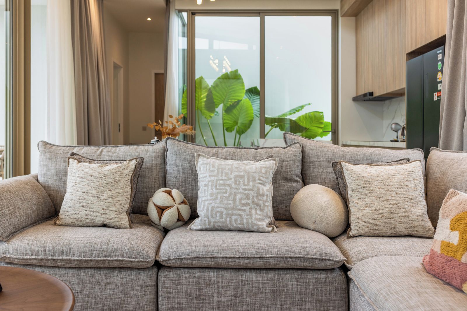 A modern living room with a grey sectional sofa, decorative pillows, a vase with green plants, and a kitchen area in the background.