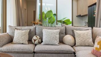 A modern living room with a grey sectional sofa, decorative pillows, a vase with green plants, and a kitchen area in the background.