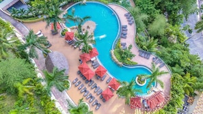 Ein großer, geschwungener Pool, umgeben von Liegestühlen, Sonnenschirmen und Palmen.