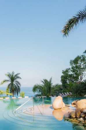 A clear pool with a metal ladder, surrounded by rocks and palm trees, overlooking a body of water.
