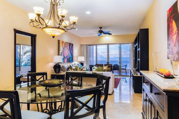 A spacious living area with a dining table, chairs, a chandelier, a ceiling fan, and a large window offering a view of the sea.