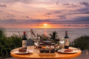 Un tavolo apparecchiato con bottiglie di vino, bicchieri e cibo, con vista su un tramonto sull'oceano.