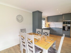 A dining area with a wooden table and white chairs, a kitchen with grey cabinets, and a wall-mounted clock. - The Willows (Mablethorpe)