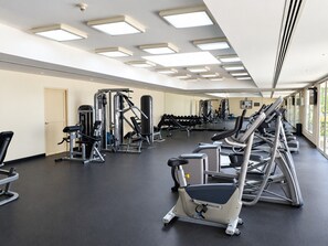 A gym with various exercise equipment, including treadmills and weight machines, and a large window providing natural light. - HAVN - Cozy Studio in Ghozlan, The Greens (Dubai)
