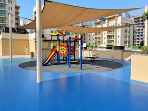 A playground with a slide and climbing structures, surrounded by a blue floor and beige buildings in the background. - HAVN - Cozy Studio in Ghozlan, The Greens (Dubai)