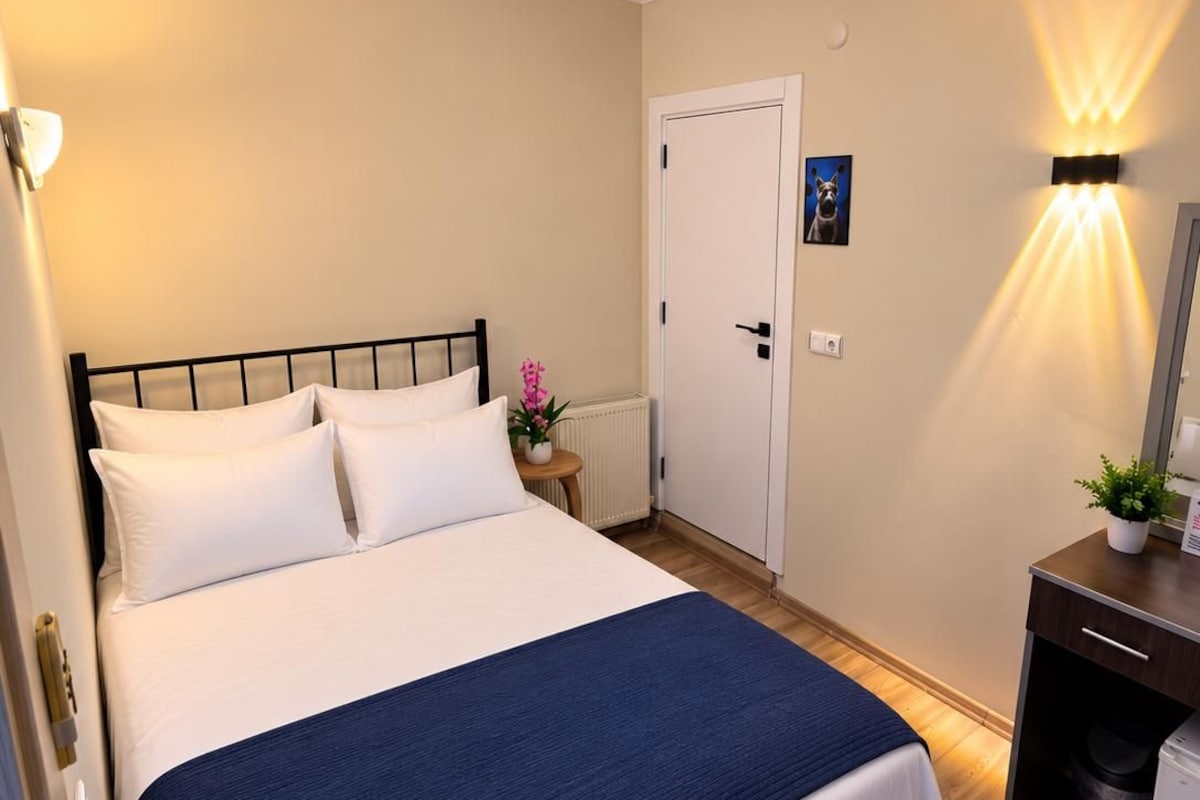 A neatly made bed with white and blue bedding, a small bedside table with a plant, a door with a dog picture, and a wall-mounted light fixture.