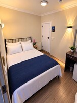 A neatly made bed with white and blue bedding, a small bedside table with a plant, a door with a dog picture, and a wall-mounted light fixture.