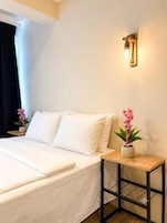 Una cama bien hecha con sábanas blancas, una mesita de noche con un jarrón de flores rosas y una lámpara de pared.