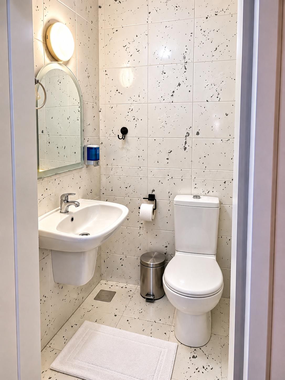 A bathroom with a sink, mirror, toilet, and a trash bin.