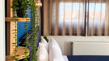 Quarto com cama, almofadas brancas, planta pendente, cabeceira de madeira e janela com cortinas.