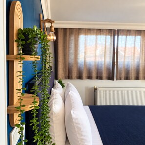 Un dormitorio con cama, almohadas blancas, una planta colgante, cabecera de madera y ventana con cortinas.