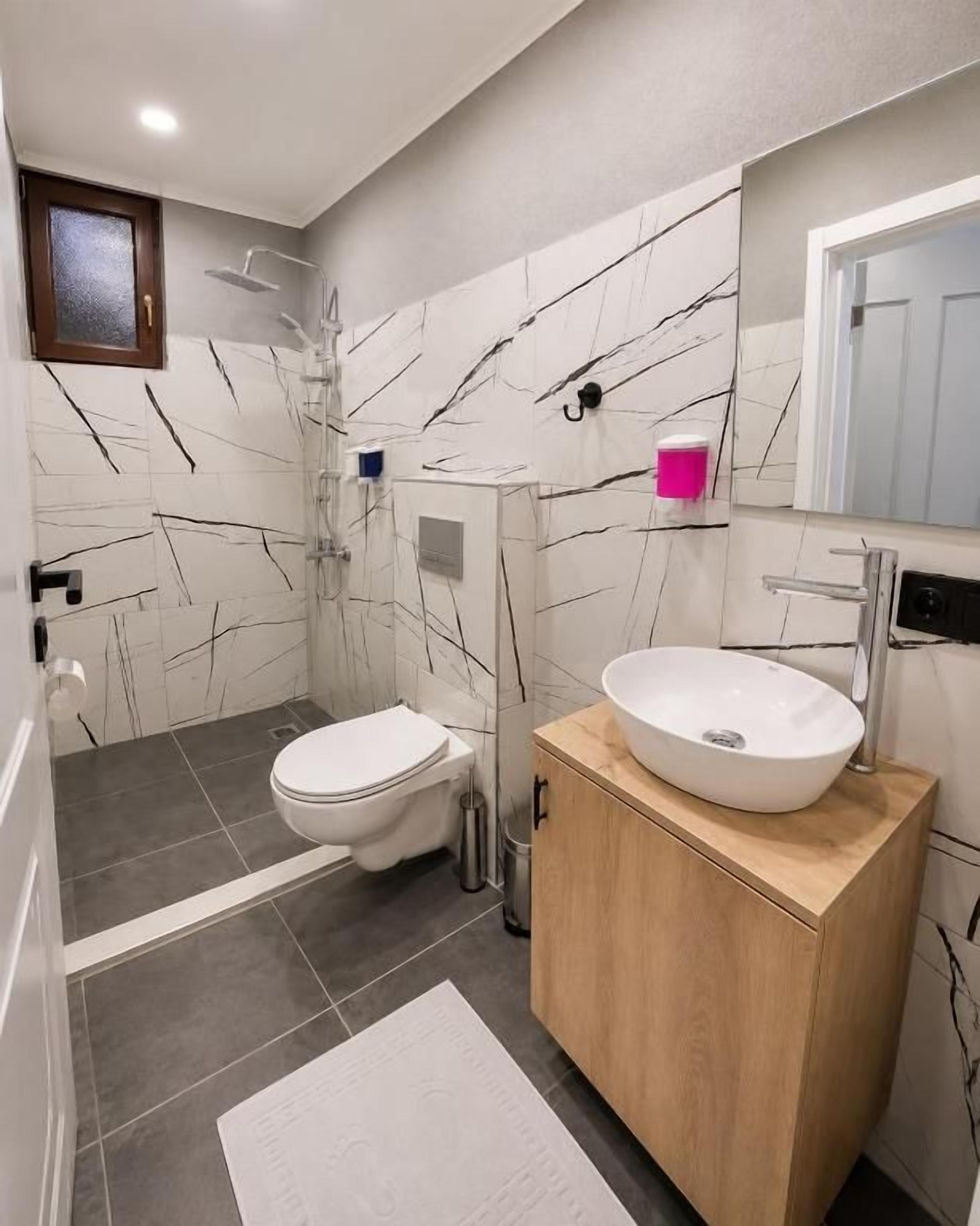 Un baño moderno con pared de mármol, tocador de madera, lavamanos blanco y un inodoro.