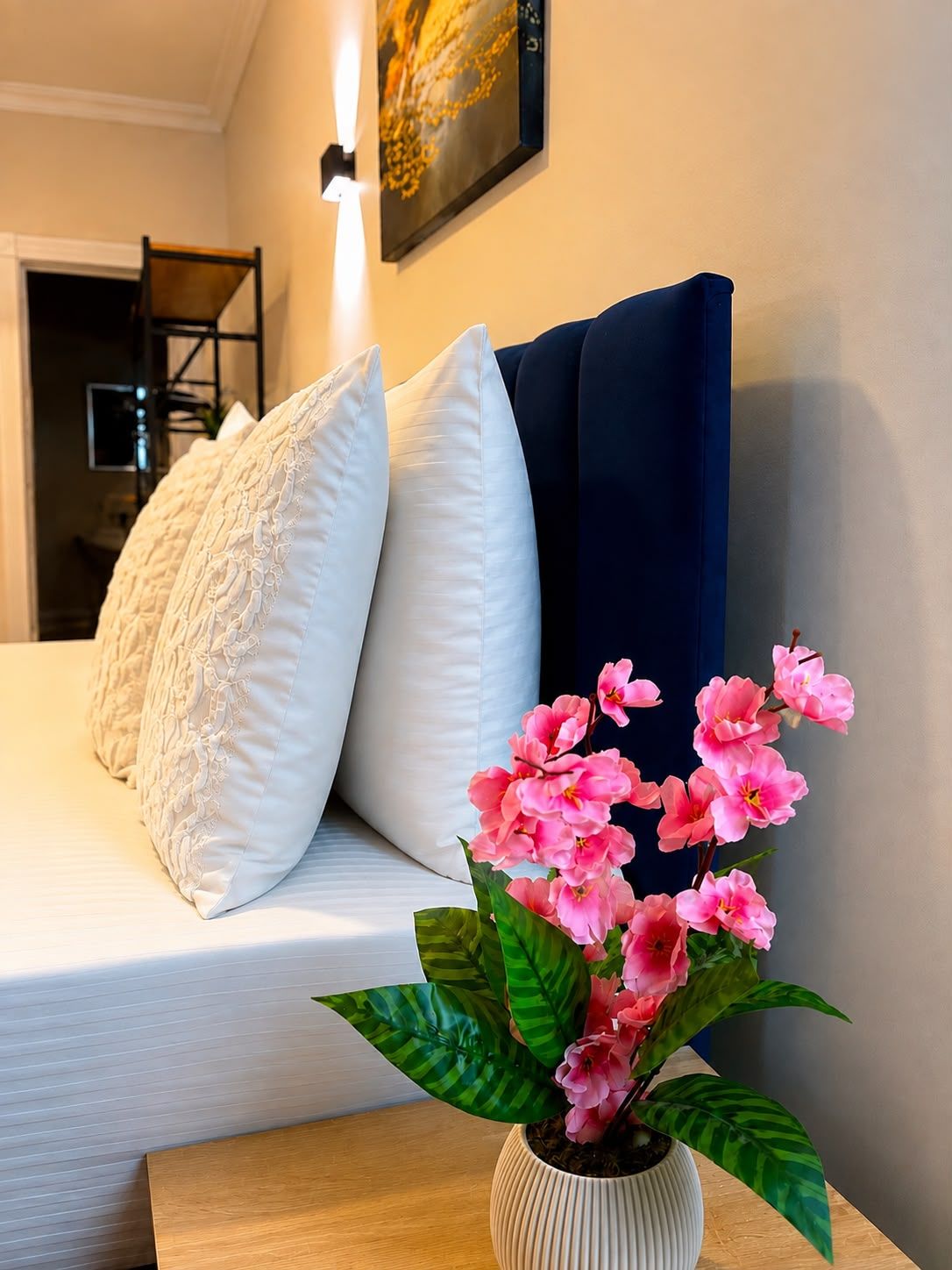 Una cama con sábanas blancas, dos almohadas, una cortina azul oscuro y un florero con flores rosadas sobre una mesita de noche de madera.