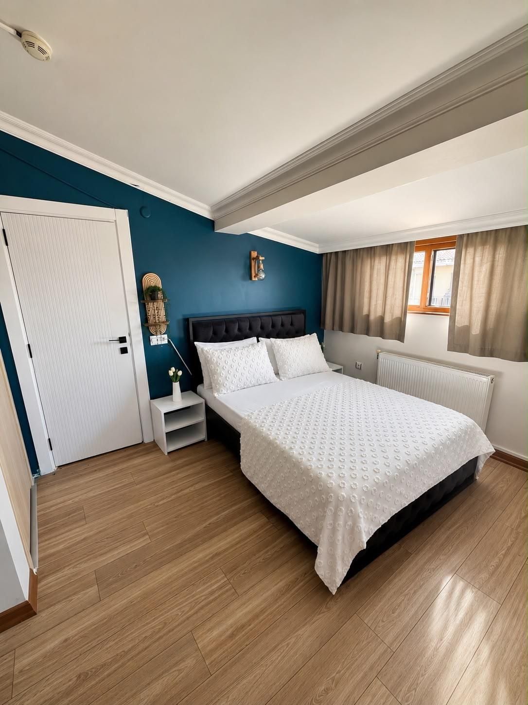 Un dormitorio con cama, una mesita de noche, una lámpara de pared y una ventana con cortinas.