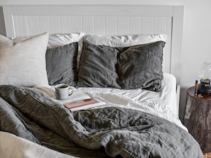 A bed with grey bedding, a grey mug, and a book on a wooden nightstand.
