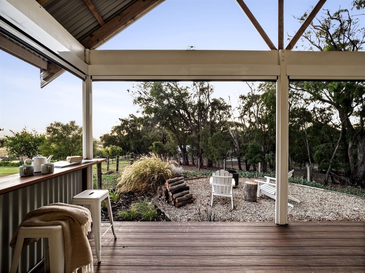 A covered outdoor area with a wooden deck, white chairs, a small table, and a view of a garden with trees and a gravel path.