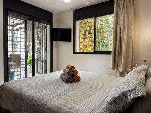 A hotel room with a bed, a TV mounted on the wall, a window with curtains, and a view of greenery outside. - Appartement Familial Spacieux – Proche Aéroport & Tram, Parking Gratuit (Salé)
