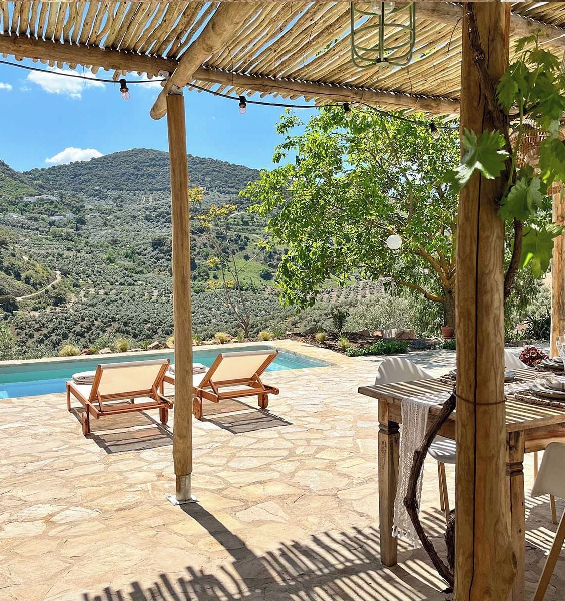 A poolside area with wooden chairs and a table, surrounded by a pergola with string lights.