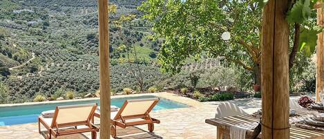 A poolside area with wooden chairs and a table, surrounded by a pergola with string lights.