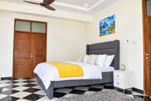 A bedroom with a bed, a nightstand, a wooden wardrobe, a ceiling fan, and a painting on the wall. - The Coastal Nest Diani Beach (Diani Beach)
