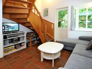 Ein Wohnzimmer mit einer Holztreppe, einem Fernseher, einem Sofa und einem Fenster mit Blick ins Grüne.