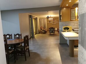 Ein Essbereich mit Holztisch und -stühlen, eine Küche mit Marmorarbeitsplatte und ein Wohnbereich mit Sofa und Vorhängen.
