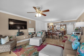 A spacious living room with a ceiling fan, a flat-screen TV, and a dining area with a chandelier. - Edgewater Beach Condo 202 (Destin)