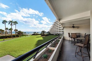 A balcony with a view of a well-maintained lawn, palm trees, and the ocean. - Edgewater Beach Condo 302 (Destin)