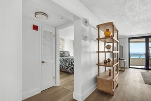 A hotel room with a wooden floor, a bed with patterned bedding, a white door, and a shelf with decorative items. - Edgewater Beach Condo 505 (Destin)