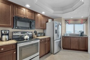 Una cocina con armarios de madera, estufa de acero inoxidable, microondas, refrigerador y una ventana con persianas.
