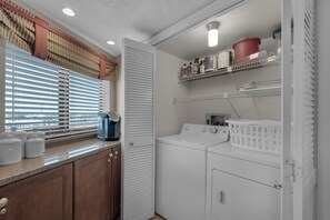 A laundry room with a washing machine, dryer, and shelves with various items. - Edgewater Beach Condo 0603 (Destin)
