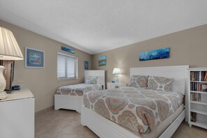 A bedroom with two beds, a bedside table, a lamp, a bookshelf, and artwork on the walls. - Edgewater Beach Condo 0603 (Destin)