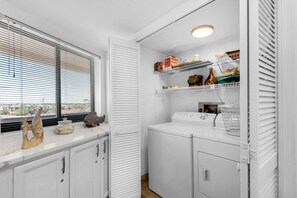 A laundry room with a washing machine, drying rack, and shelves with various items. - Edgewater Beach Condo 608 (Destin)