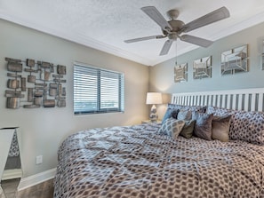 Ein Schlafzimmer mit Bett, Deckenventilator, Fenster mit Jalousien, Wandbild und einer Lampe.