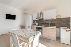 Una cucina compatta con zona pranzo, mobili bianchi, una TV a schermo piatto e un piccolo tavolo con tovaglia floreale.