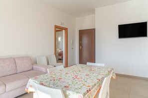 A room with a sofa, a table with a floral tablecloth, a TV mounted on the wall, and a door. - Apartment 'Lungomare Avola  with Sea' with Balcony, Wi-Fi and Air Conditioning (Avola)