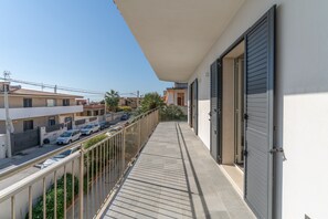 Ein Balkon mit Metallgeländer, Fliesenboden und Blick auf geparkte Autos sowie Gebäude im Hintergrund.