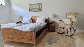 Ett sovrum med en säng i trä, två sängbord med lampor, en gungstol, en tavla på väggen och en mönstrad matta på golvet.