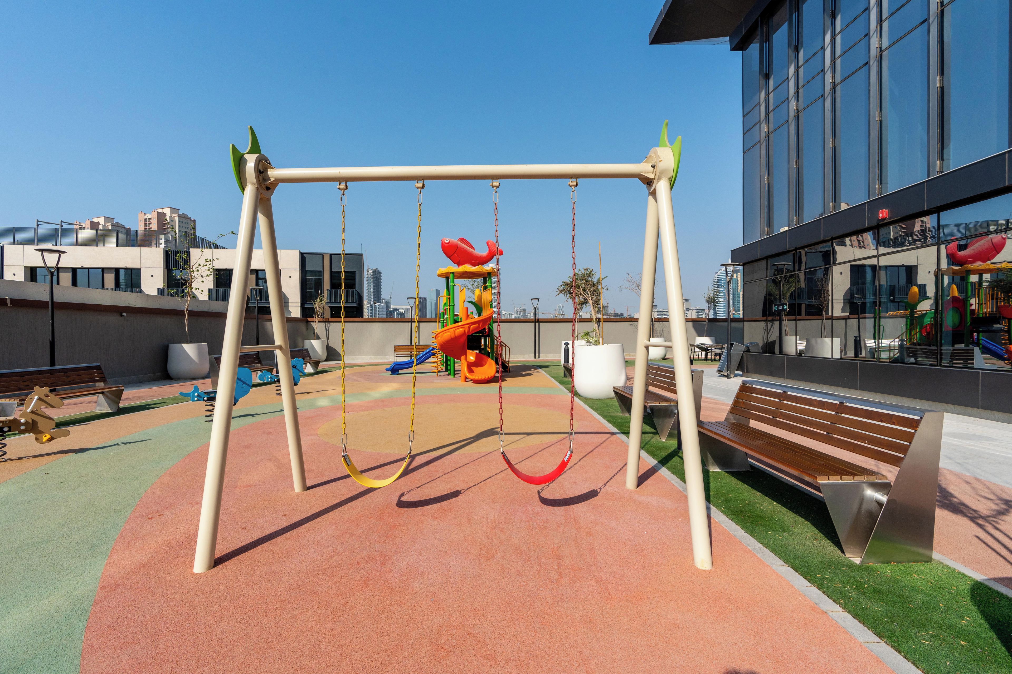 Un parque infantil con columpios, toboganes, bancos y un edificio con grandes ventanales al fondo.