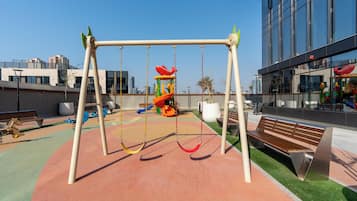 Un parque infantil con columpios, toboganes, bancos y un edificio con grandes ventanales al fondo.