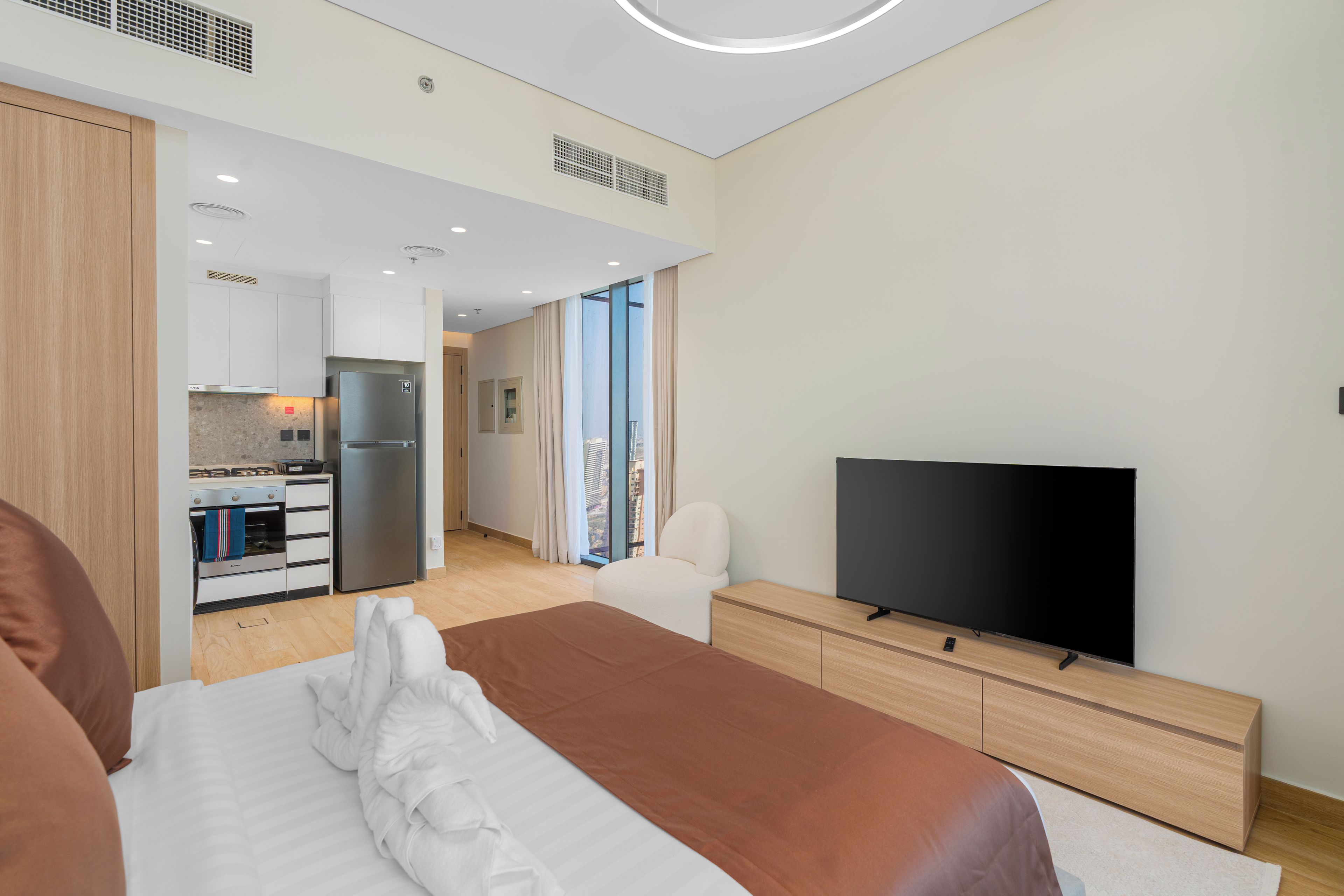Habitación de hotel moderna con cama, televisor de pantalla plana, cómoda de madera y una zona de cocina con refrigerador.
