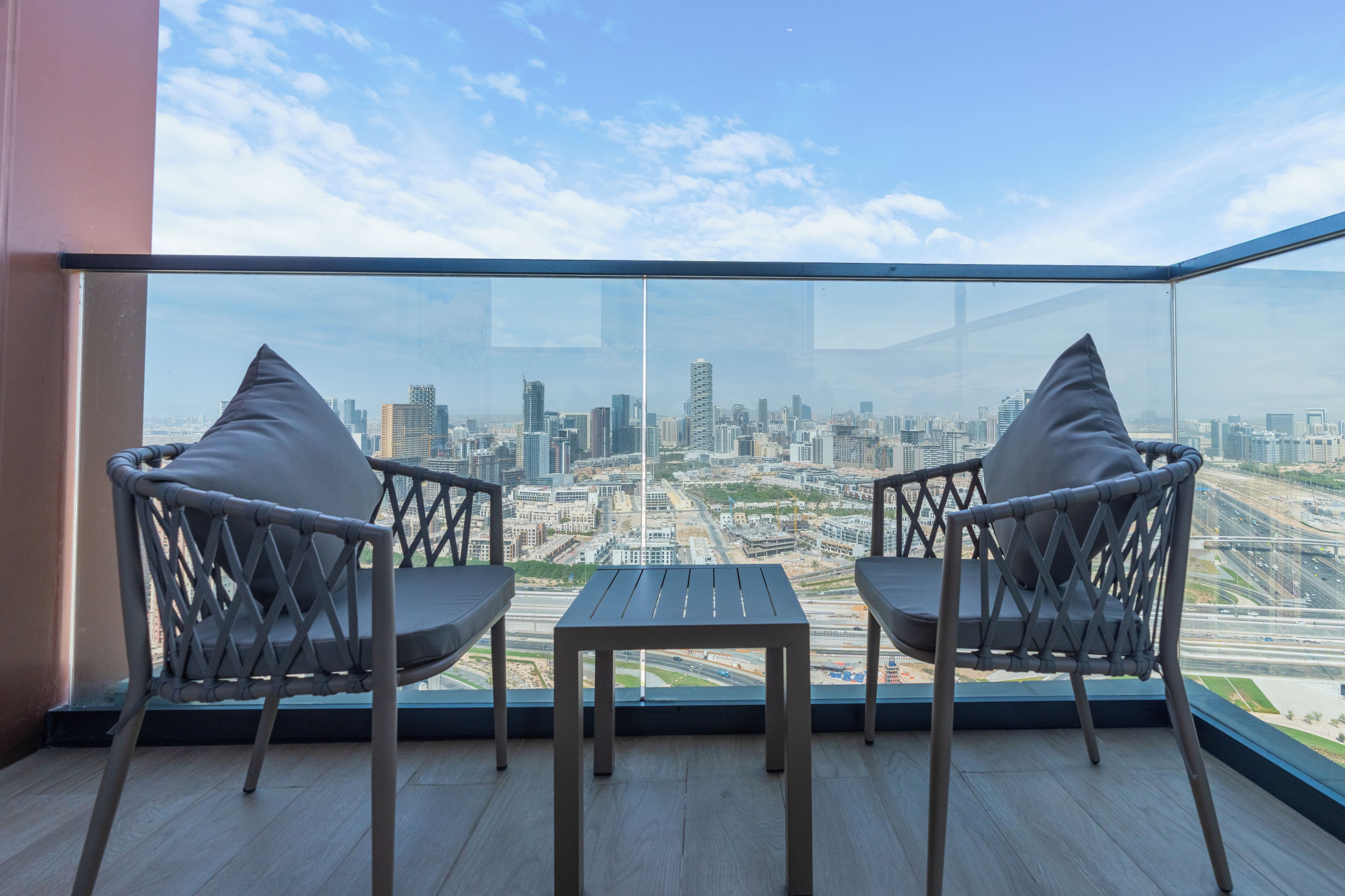 A balcony with two chairs and a small table, overlooking a cityscape with buildings and a highway.
