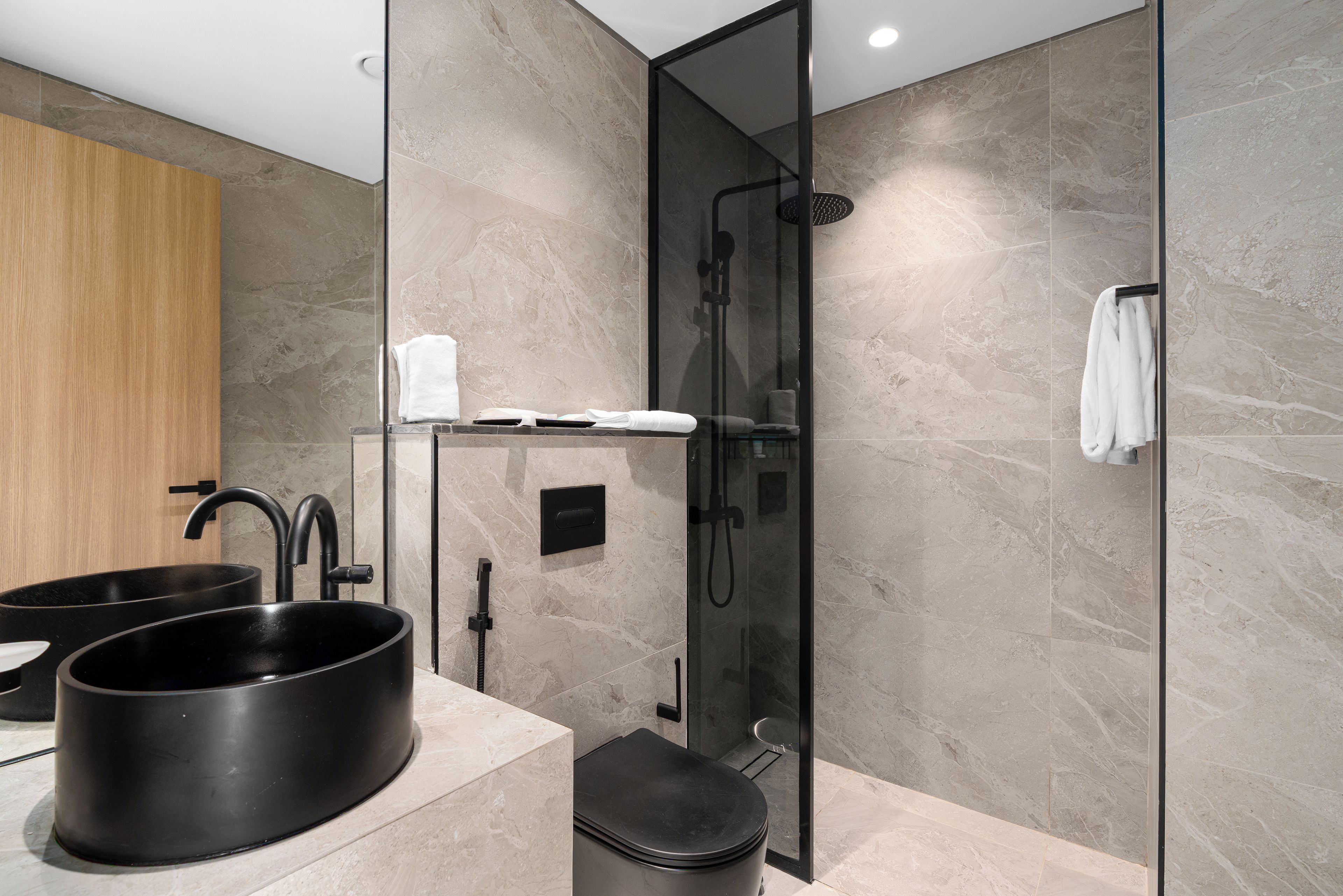 Un baño moderno con ducha envuelta en vidrio, un lavamanos negro y un mueble de tocador de madera.
