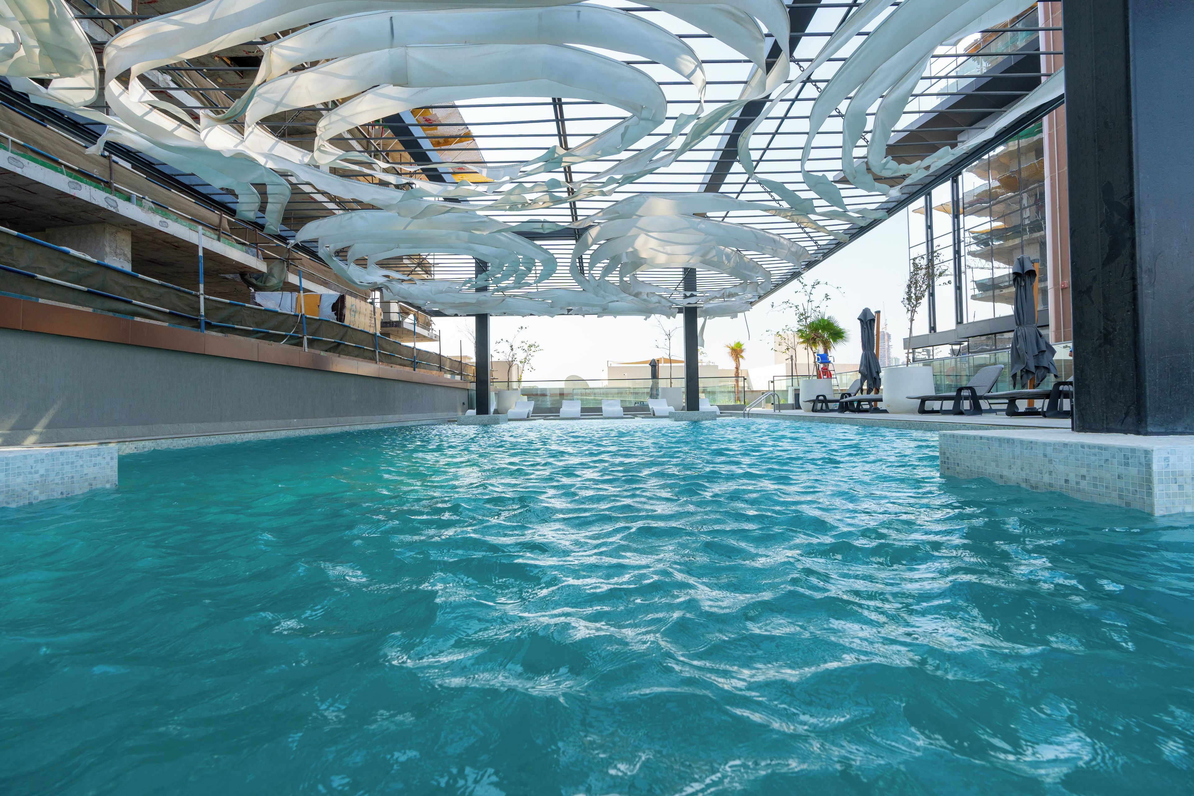 An indoor swimming pool with a modern ceiling design, surrounded by a poolside area with seating and a view of the cityscape.