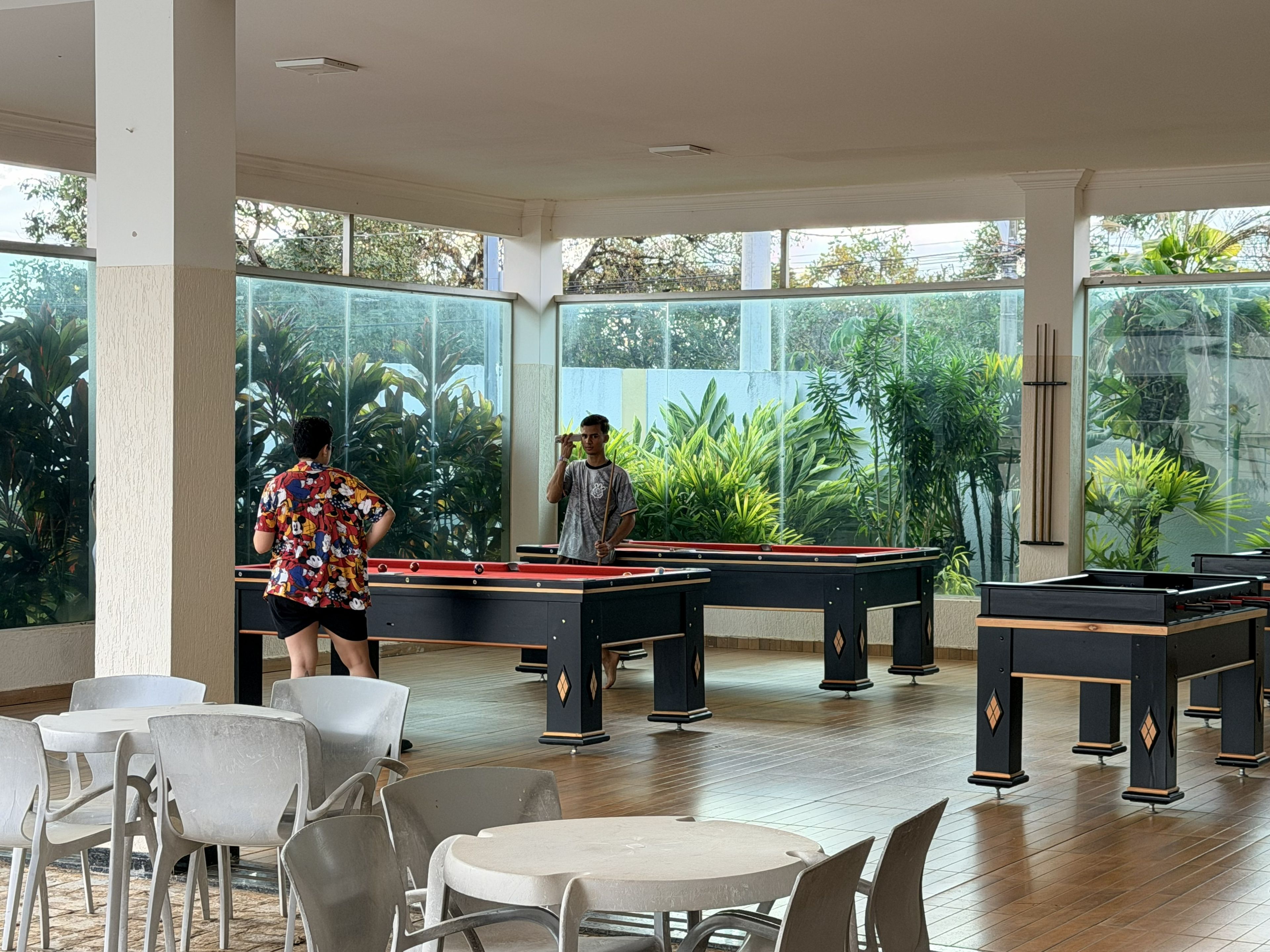 A pool table area with tables and chairs, surrounded by large windows with a view of greenery.