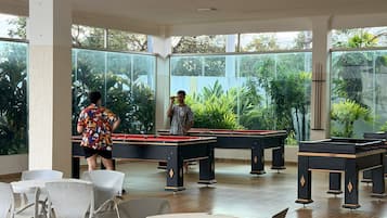A pool table area with tables and chairs, surrounded by large windows with a view of greenery.